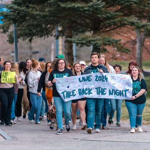 A group of students marches on campus holding a sign that reads "51СƳ2024 Take Back the Night"